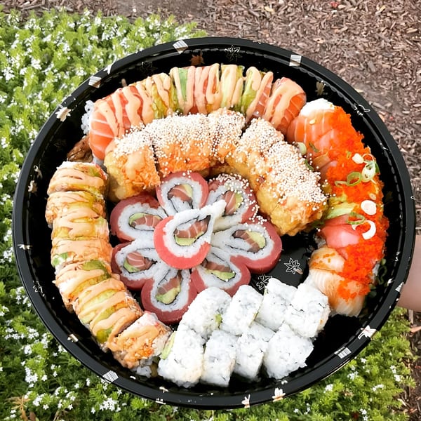 A large black platter of assorted Akita Sushi rolls, including salmon, tuna, and avocado, on a green plant background.